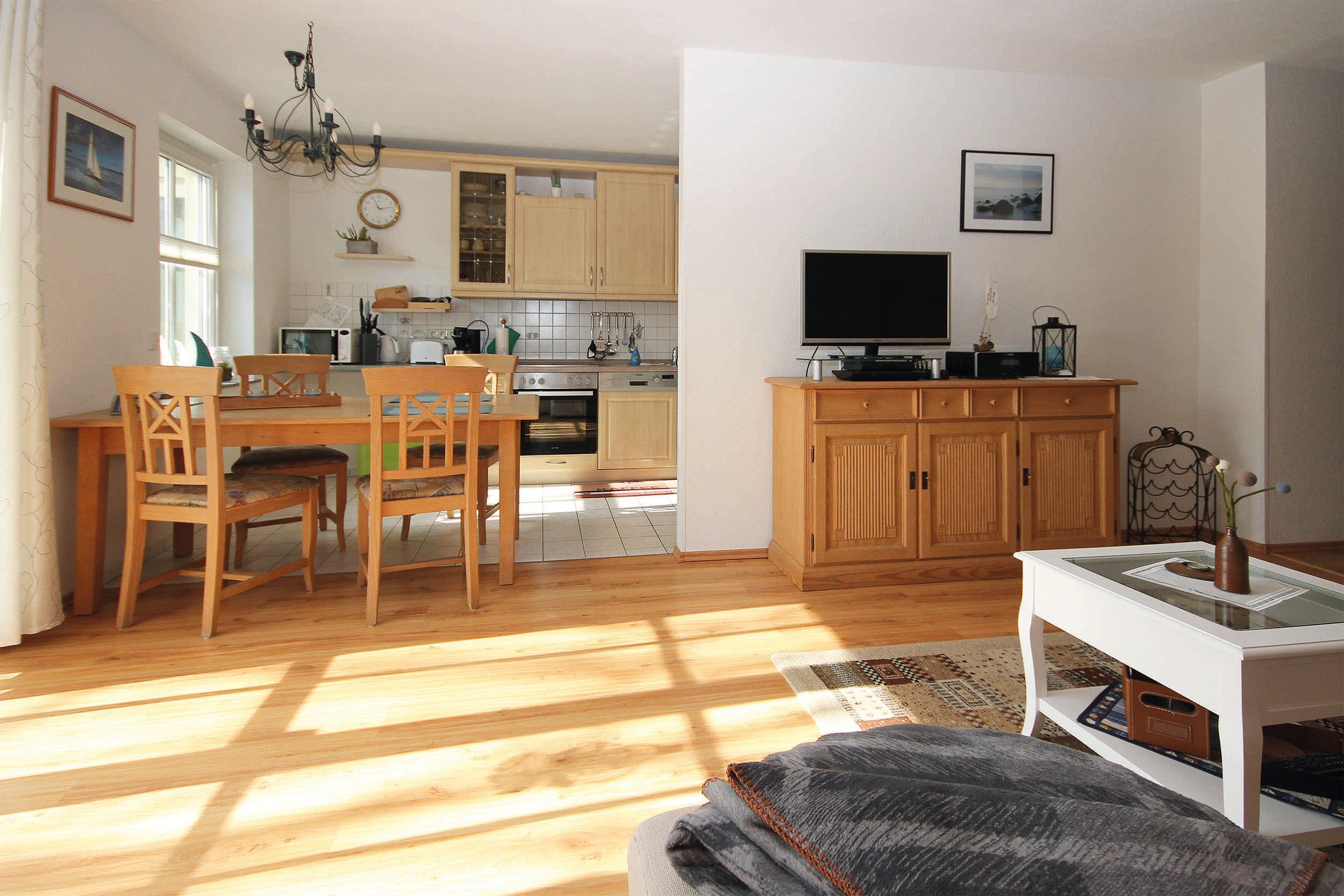 A dining area with wooden chairs and a table, a kitchen with wooden cabinets, a television on a cabinet, and a window with curtains.