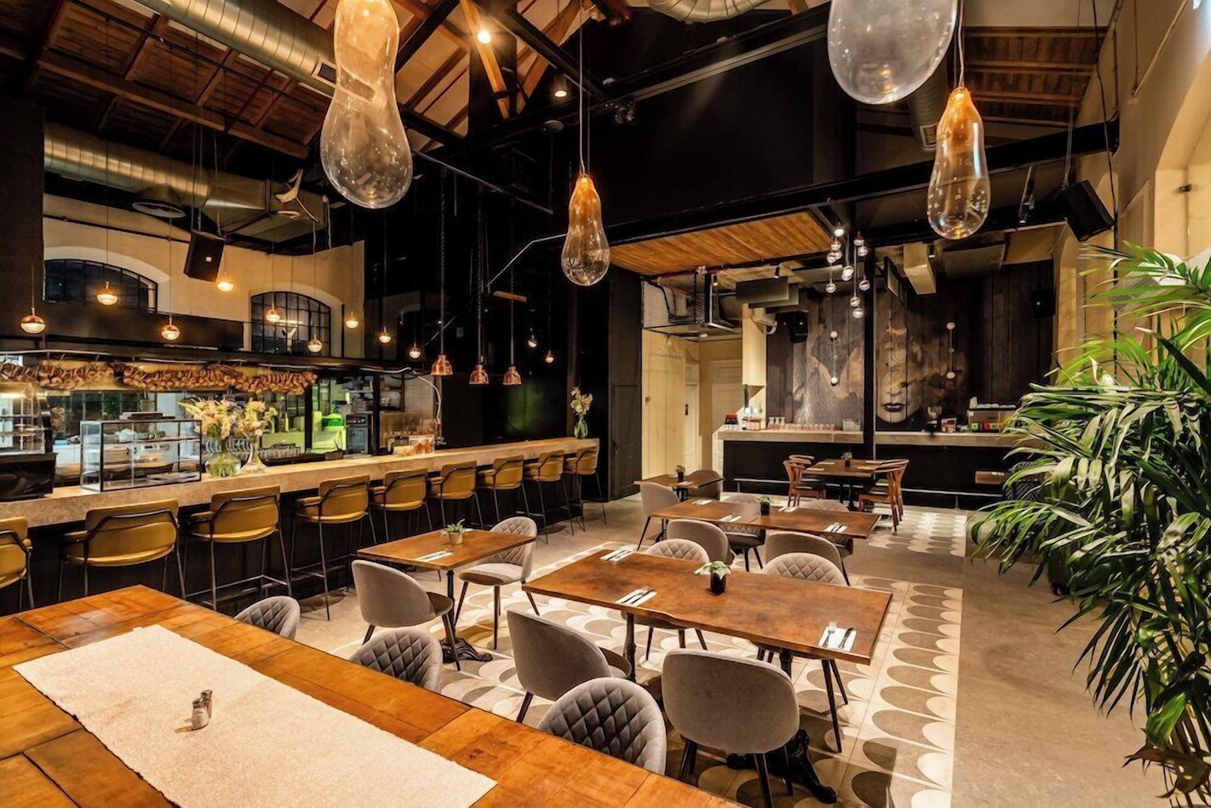 A restaurant with wooden tables and chairs, hanging pendant lights, and a bar area.