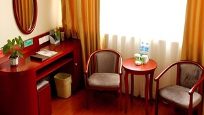 Une chambre d’hôtel équipée d’un bureau, de deux chaises, d’une petite table avec une carafe et des verres, d’une plante en pot et d’une fenêtre avec des rideaux.