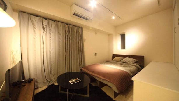 A hotel room with a bed, a small round table, a desk with a chair, and an air conditioning unit.
