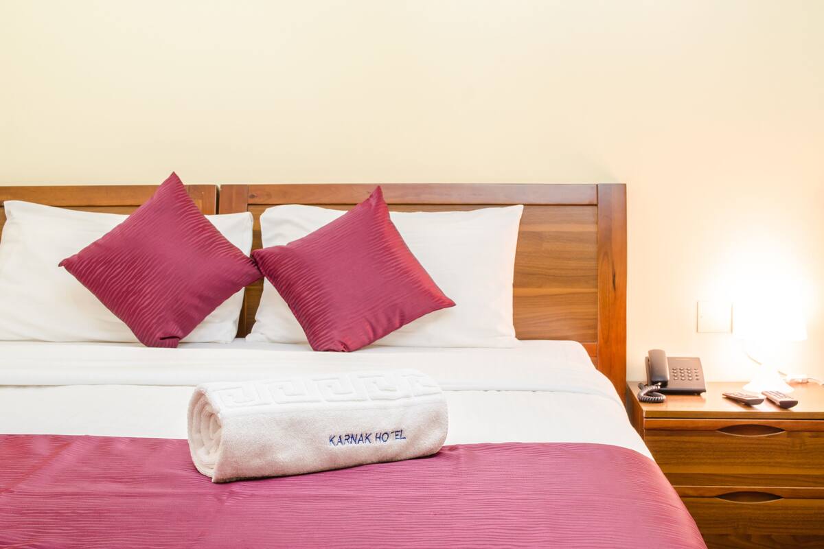Camera d'albergo con un letto rifatto, due cuscini rossi, un asciugamano piegato con la scritta 'Karnak Hotel', un comodino in legno con telefono e telecomando.