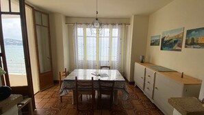 Een eethoek met een tafel en stoelen, een raam met gordijnen, een hanglamp en uitzicht op zee.