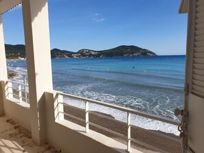 Une vue sur la côte depuis un balcon doté d’une balustrade, avec une mer bleue et limpide, et une colline lointaine parsemée de bâtiments.