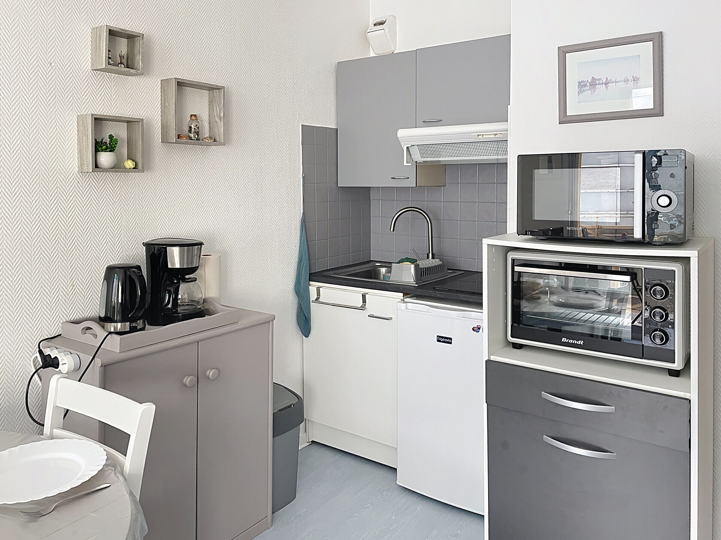 A compact kitchen with a coffee maker, toaster, and microwave. There is a sink with a faucet, a small trash bin, and a chair with a plate.