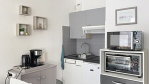 Una cucina compatta con macchina da caffè, tostapane e microonde. Si vede un lavello con rubinetto, un piccolo cestino e una sedia con un piatto.