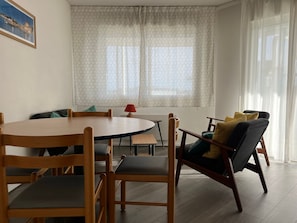 Un área de comedor con una mesa redonda de madera y sillas, una mesita auxiliar con una lámpara, y una ventana con cortinas estampadas.