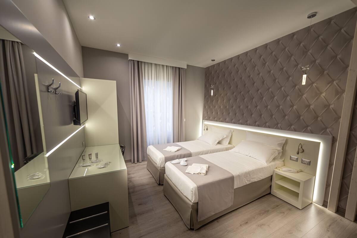 Quarto de hotel moderno com duas camas, um espelho grande, uma toucador com espelho e uma mesa de cabeceira com telefone.
