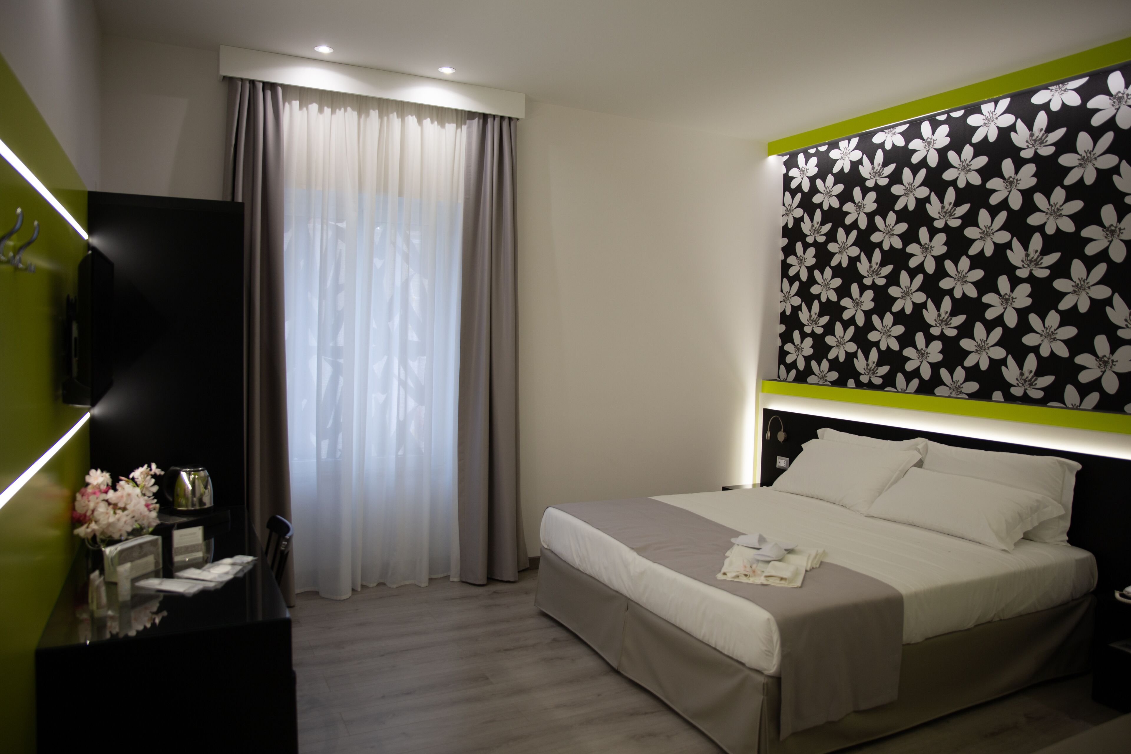 Quarto de hotel moderno com cama, televisão, escrivaninha com candeeiro e janela com cortinas.