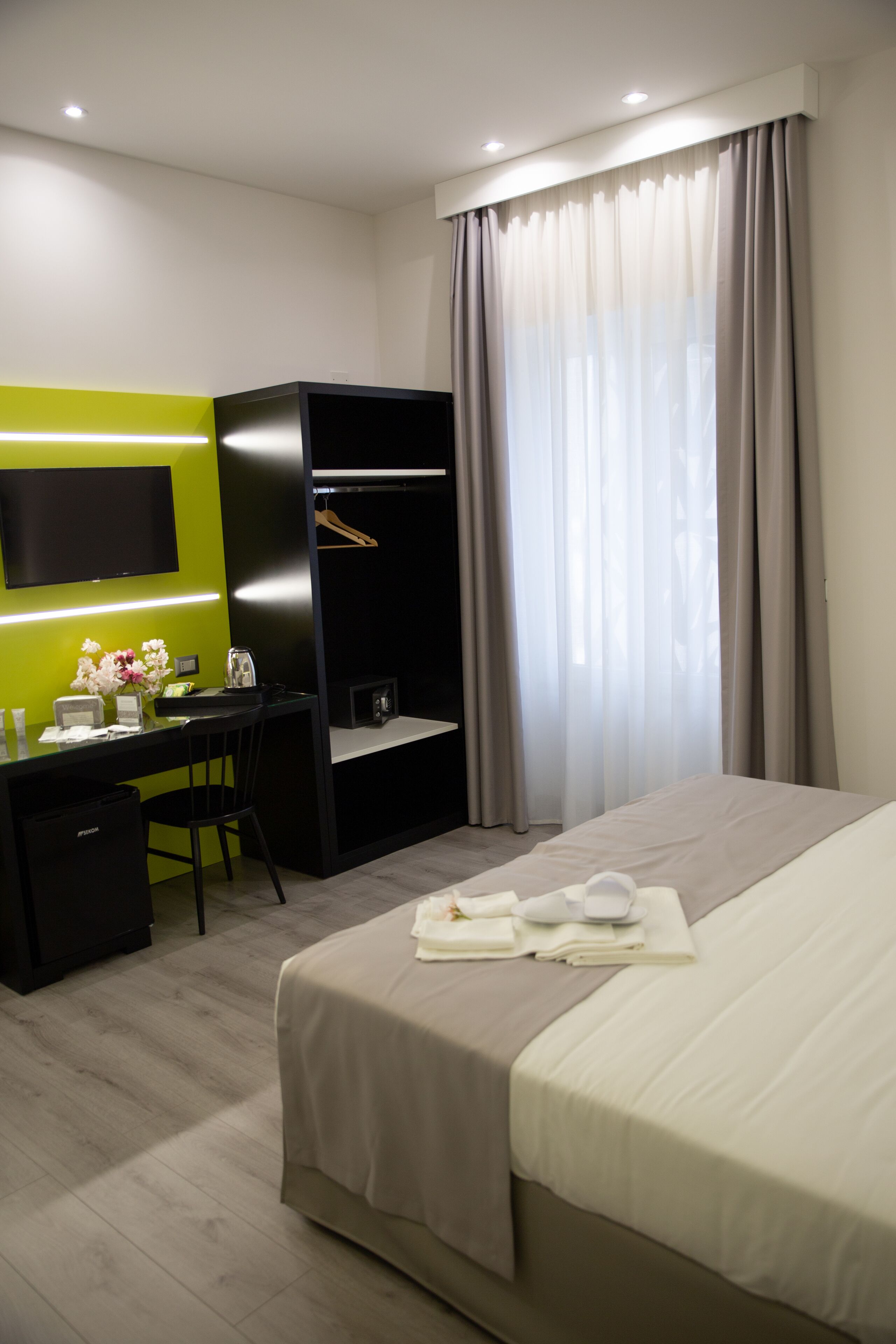 Habitación de hotel con cama, escritorio, armario, televisor y ventana con cortinas.