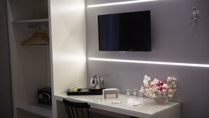 A modern hotel room with a glass desk, a flat-screen TV, a coffee machine, a welcome sign, and a vase of flowers.