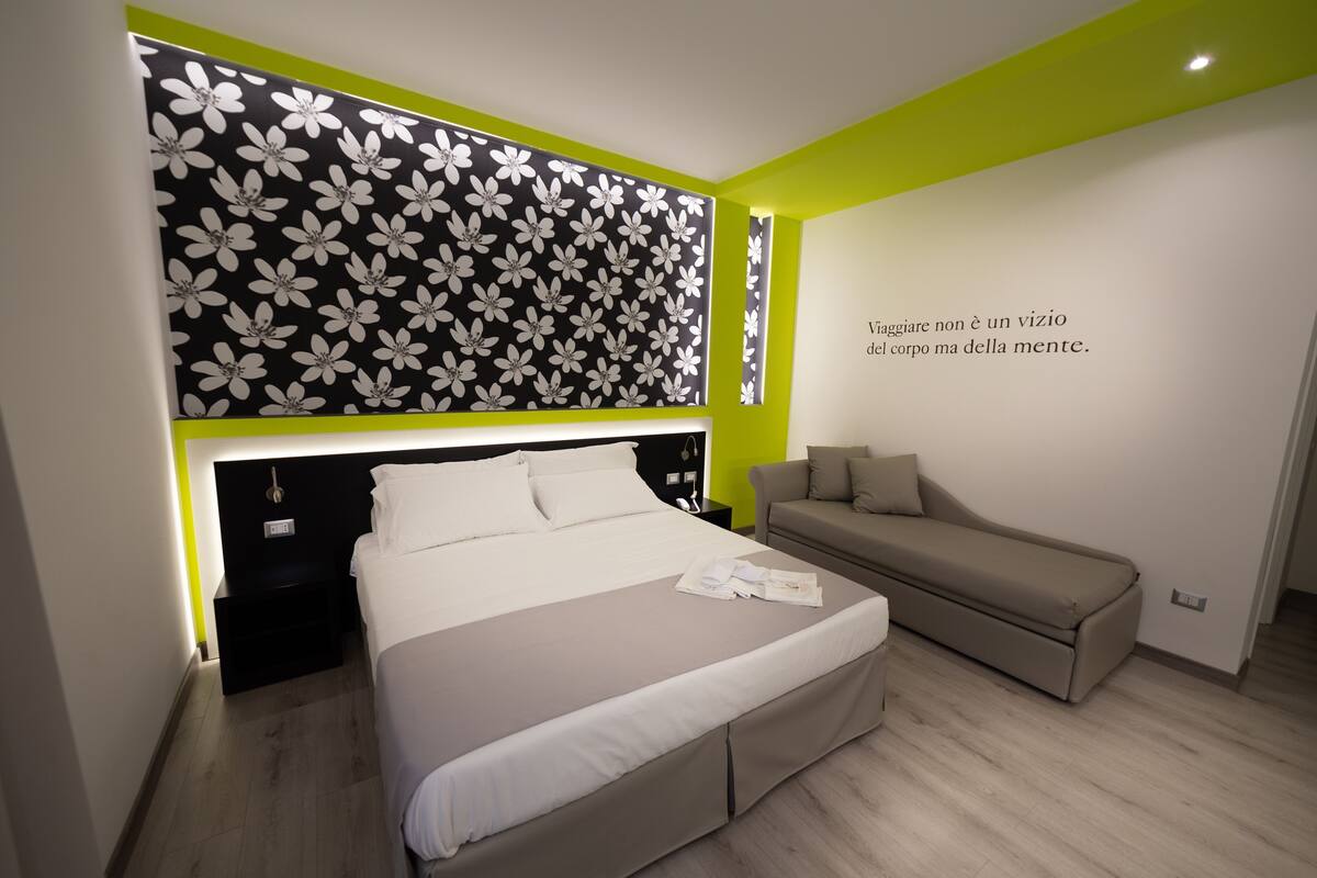 Quarto moderno com cama, sofá, parede com padrão floral e uma frase motivacional.