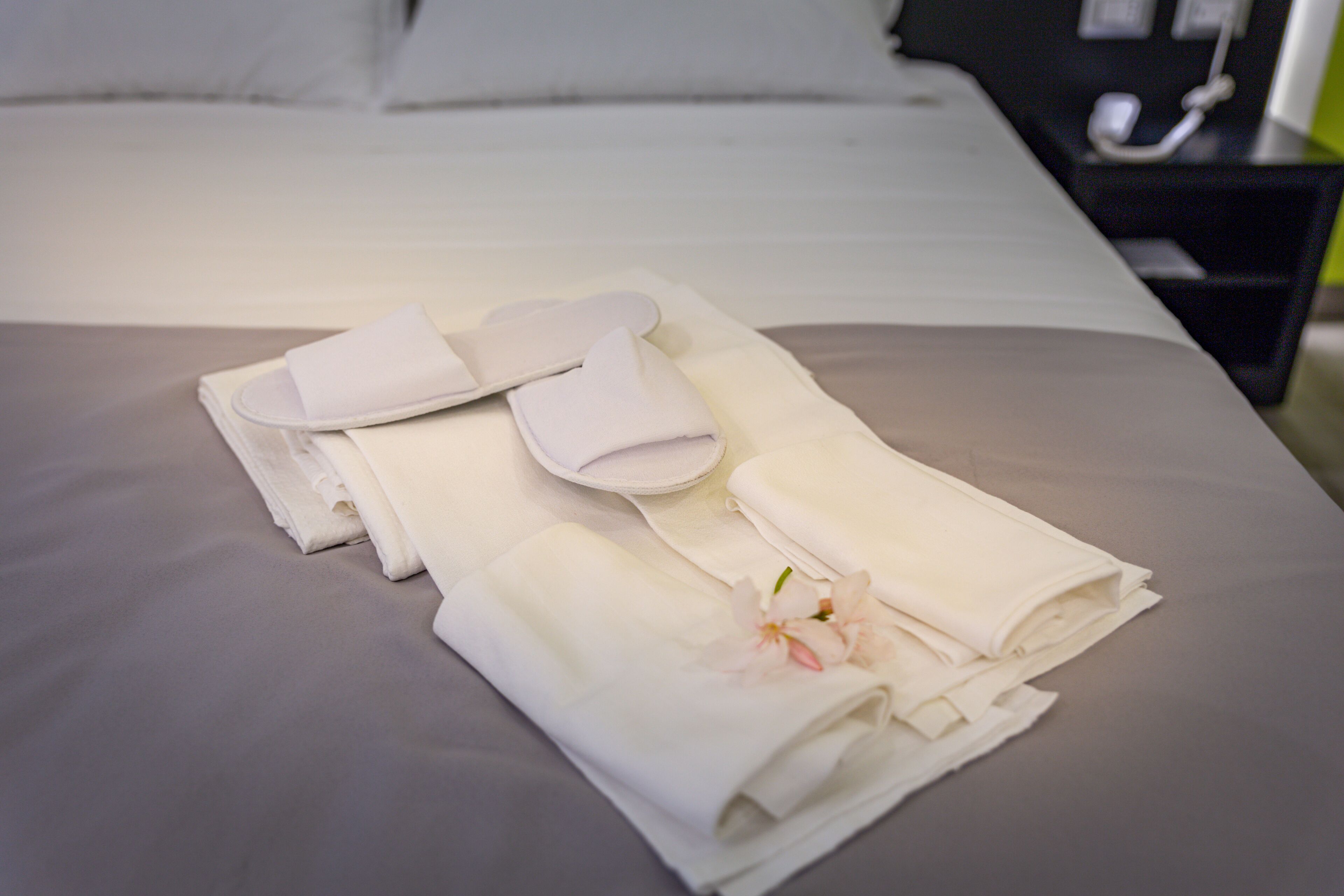 A neatly made bed with folded towels and slippers, a flower, and a bedside table with a phone and a small black object.