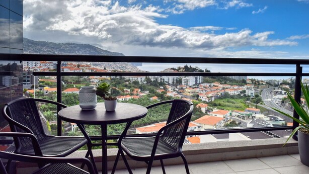 A balcony with a table and chairs, overlooking a cityscape with buildings and a mountain in the distance. - Design Gardens, a Home in Madeira (Funchal)