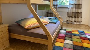 Un letto a castello con struttura in legno, un comodino con cassetto, un tappeto colorato e una finestra con tende.