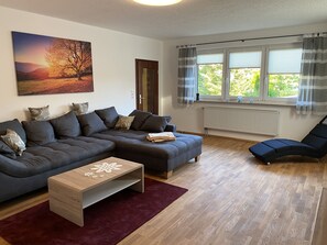 Een moderne woonkamer met een hoekbank, een klein houten bijzettafeltje en een fauteuil bij een raam met gordijnen.