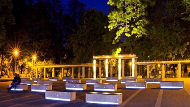 Ein Außenbereich mit Bänken und einer Pergola, umgeben von Bäumen und Straßenlaternen.