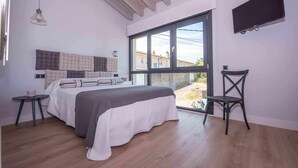 Ein Schlafzimmer mit großem Fenster, einem Bett mit grau-weißer Bettdecke, einem Nachttisch und einem Sessel.