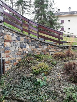 Mural de suporte de pedra com cerca de madeira, rodeada por vegetação e um edifício ao fundo.