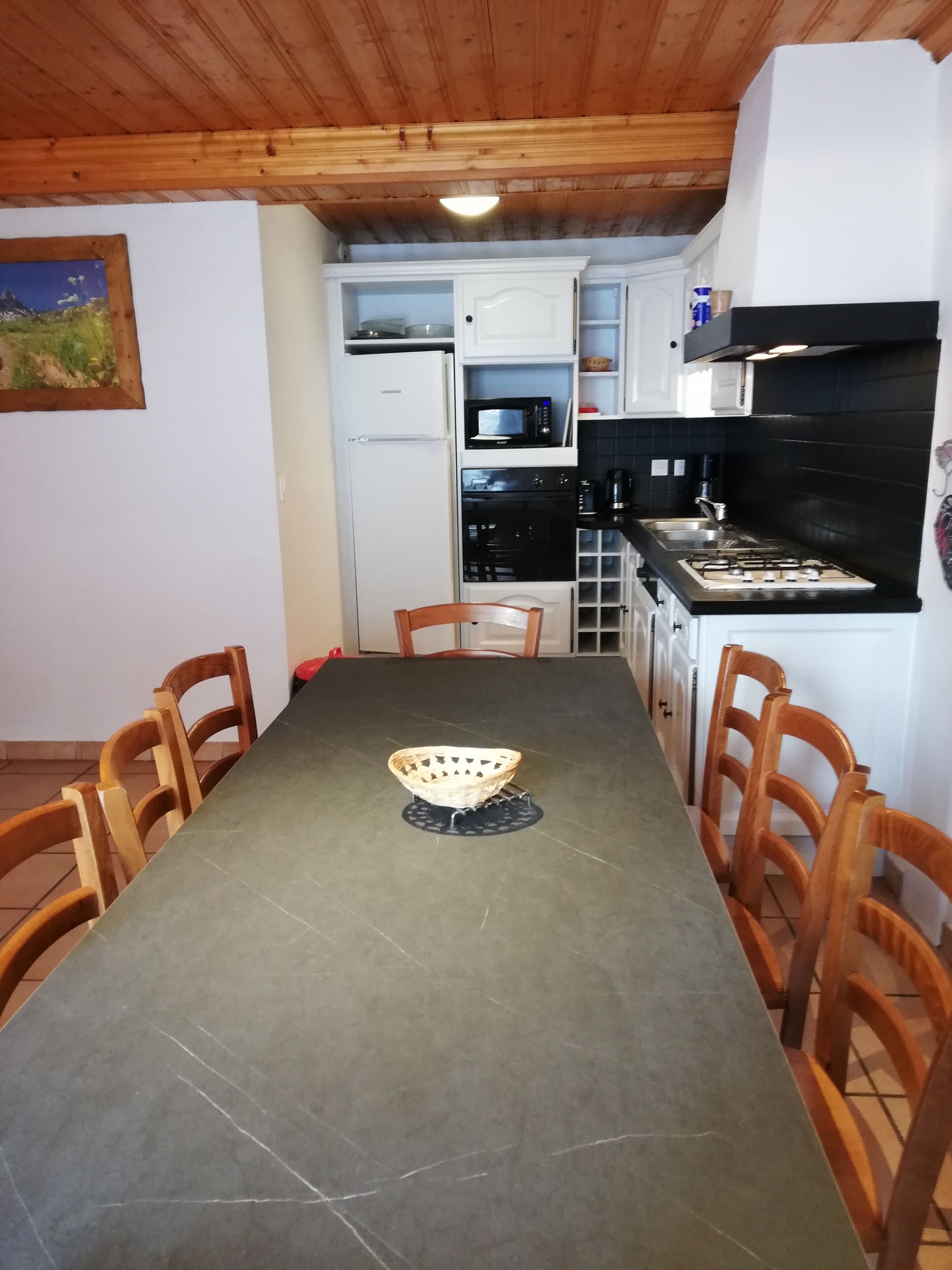 Ruokailutila, jossa on pöytä, tuoleja, kori sekä keittiö, jossa on kodinkoneet.