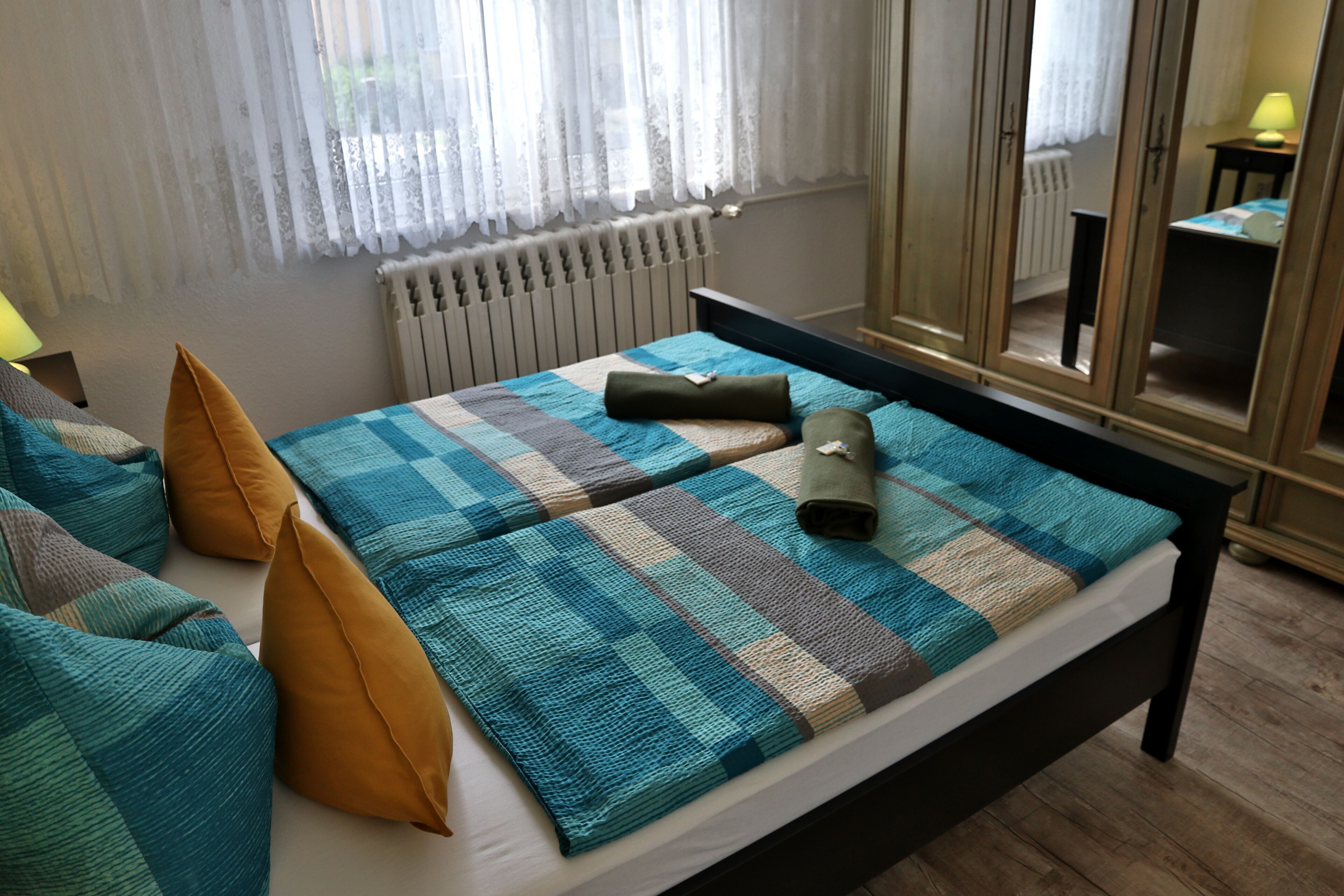 Un lit bien fait, avec une couverture à carreaux et des oreillers, un radiateur, un miroir et une lampe sur une table de chevet.