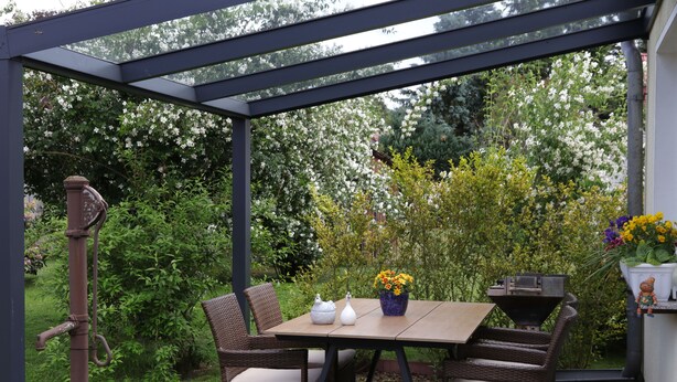 Eine überdachte Terrasse mit Tisch und Stühlen, umgeben von üppiger Bepflanzung und blühenden Pflanzen.