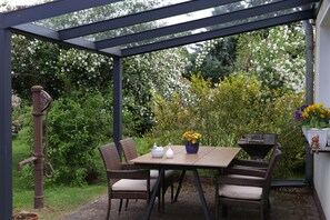 Une terrasse couverte, agrémentée d’une table et de chaises, entourée d’une végétation luxuriante et de plantes en fleurs.