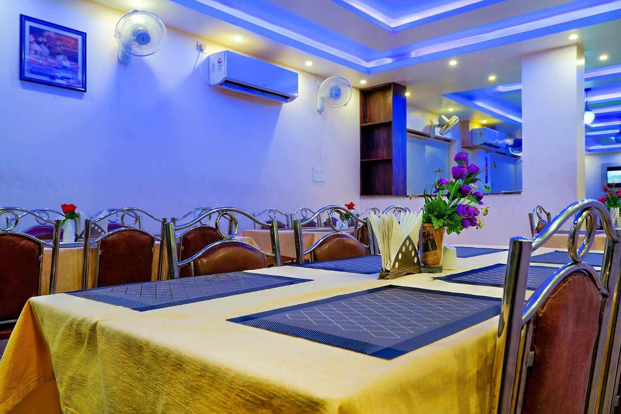 Un espace repas avec des tables dressées pour quatre personnes, des chaises et un vase rempli de fleurs violettes.