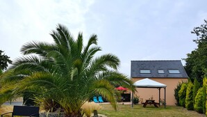 A garden area with a bench, a large palm tree, a covered picnic table, and a building with a sloped roof. - House with sauna. Internet, Comfortable, Beaches 5mn. Golf 2mn (Lamballe Armor)