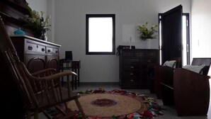 A living room with a wooden rocking chair, a sofa, a coffee table, and a colorful rug. There is a window with blinds, a cabinet, and a door.