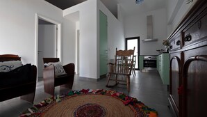 A modern living room with a bed, a wooden rocking chair, and a colorful woven rug. A kitchen with a stove and cabinets is visible in the background.