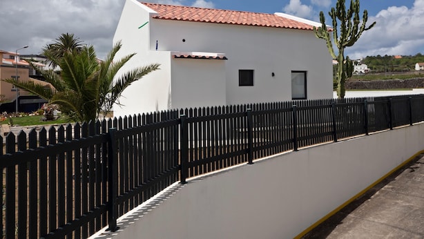 A white building with a red-tiled roof, a black fence, and a palm tree in the foreground.