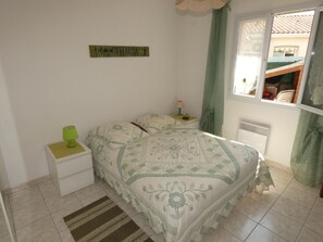 A bedroom with a bed, a nightstand, a lamp, a window with curtains, and a radiator. - Single-storey house between beaches and shops (Vaux-sur-Mer)