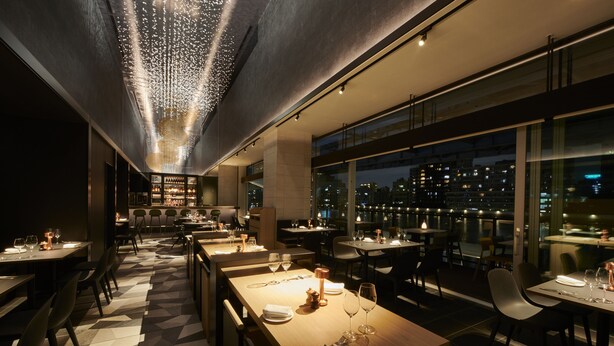 Un restaurante moderno con un suelo de patrón geométrico, mesas dispuestas con copas de vino y cubiertos, y vistas a la ciudad a través de amplios ventanales.