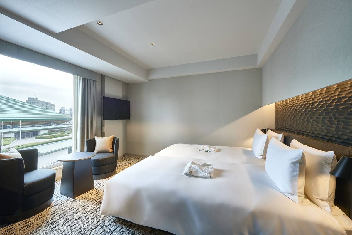A hotel room with a large bed, two armchairs, a small table, a TV, and a city view.