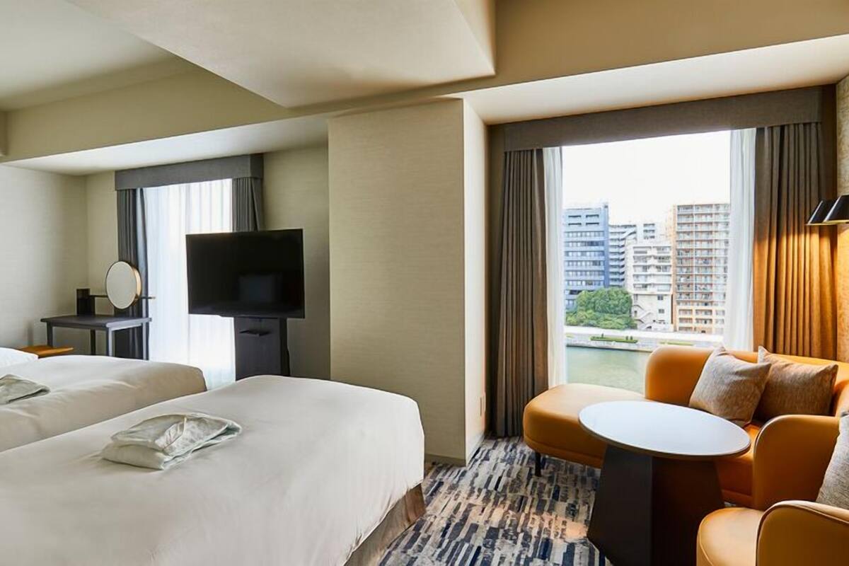 A hotel room with two beds, a TV, a small table, and a view of buildings.