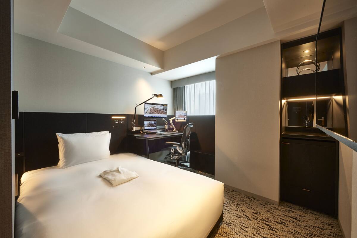 A hotel room with a large bed, a desk with a computer, a chair, and a small kitchenette.