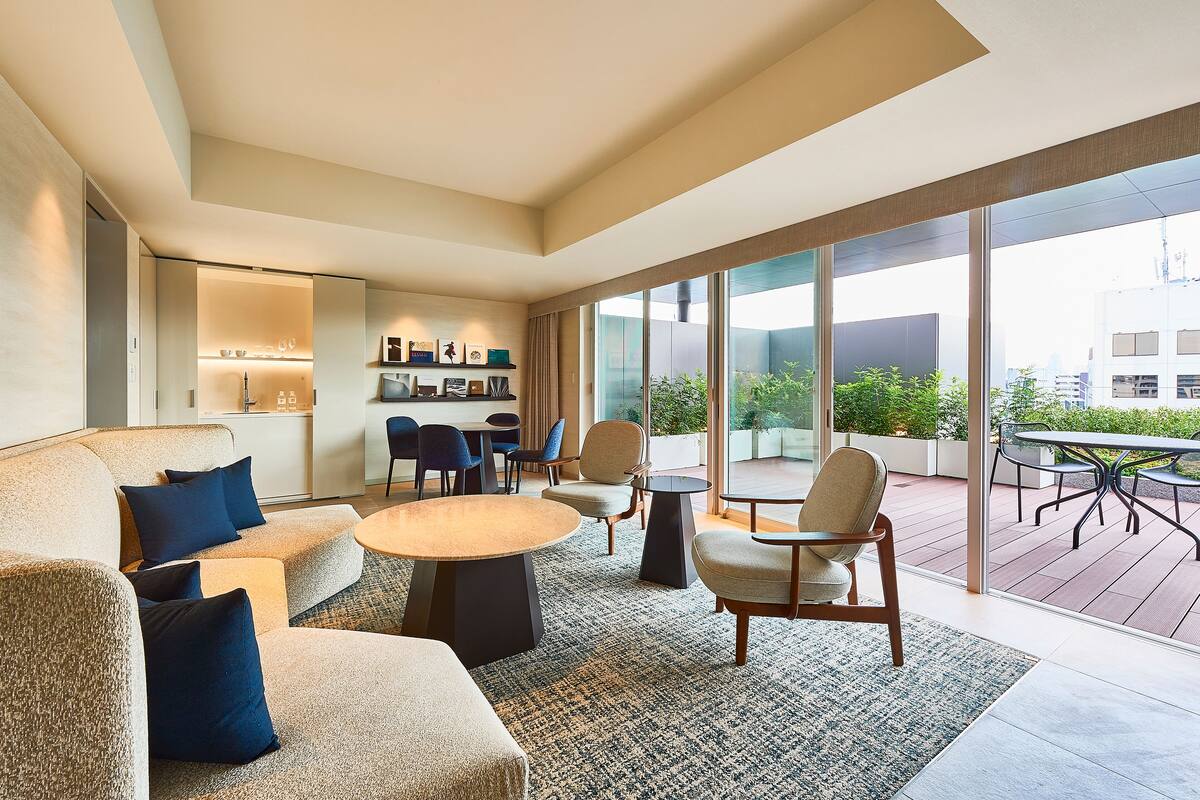 A modern living room with a sofa, chairs, a coffee table, and a dining area with a table and chairs. There is a large window providing a view of the outdoors.