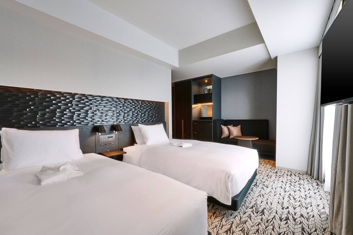 A hotel room with two beds, a modern headboard, a small seating area, and a TV.