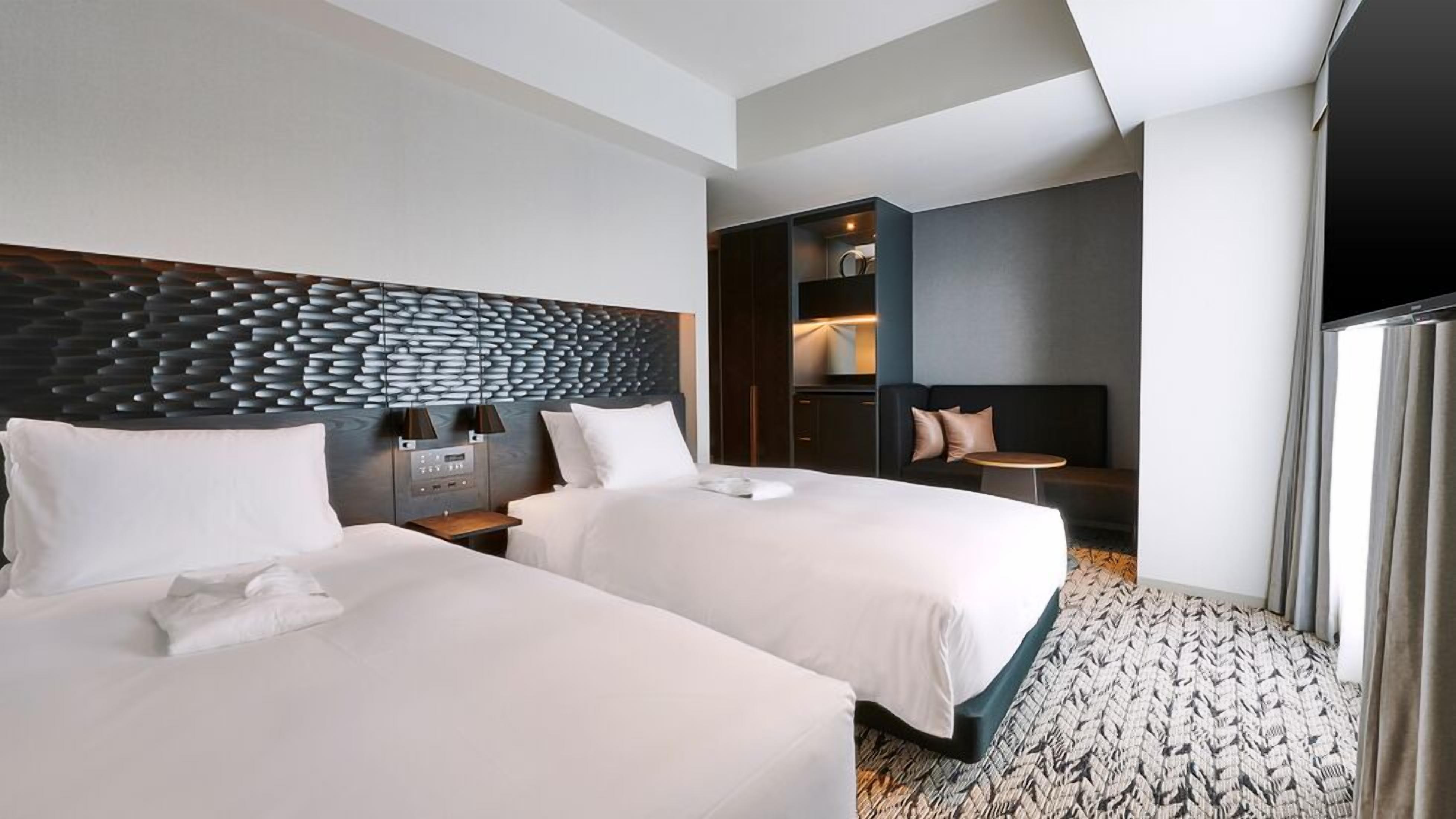 Een hotelkamer met twee bedden, een modern hoofdbord, een tafeltje en een zithoek met een bank en een fauteuil.