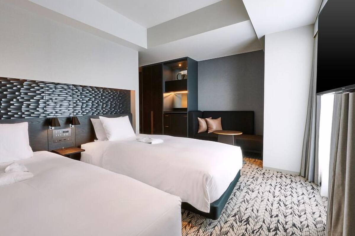 A hotel room with two beds, a modern headboard, a small table, and a seating area with a sofa and a chair.