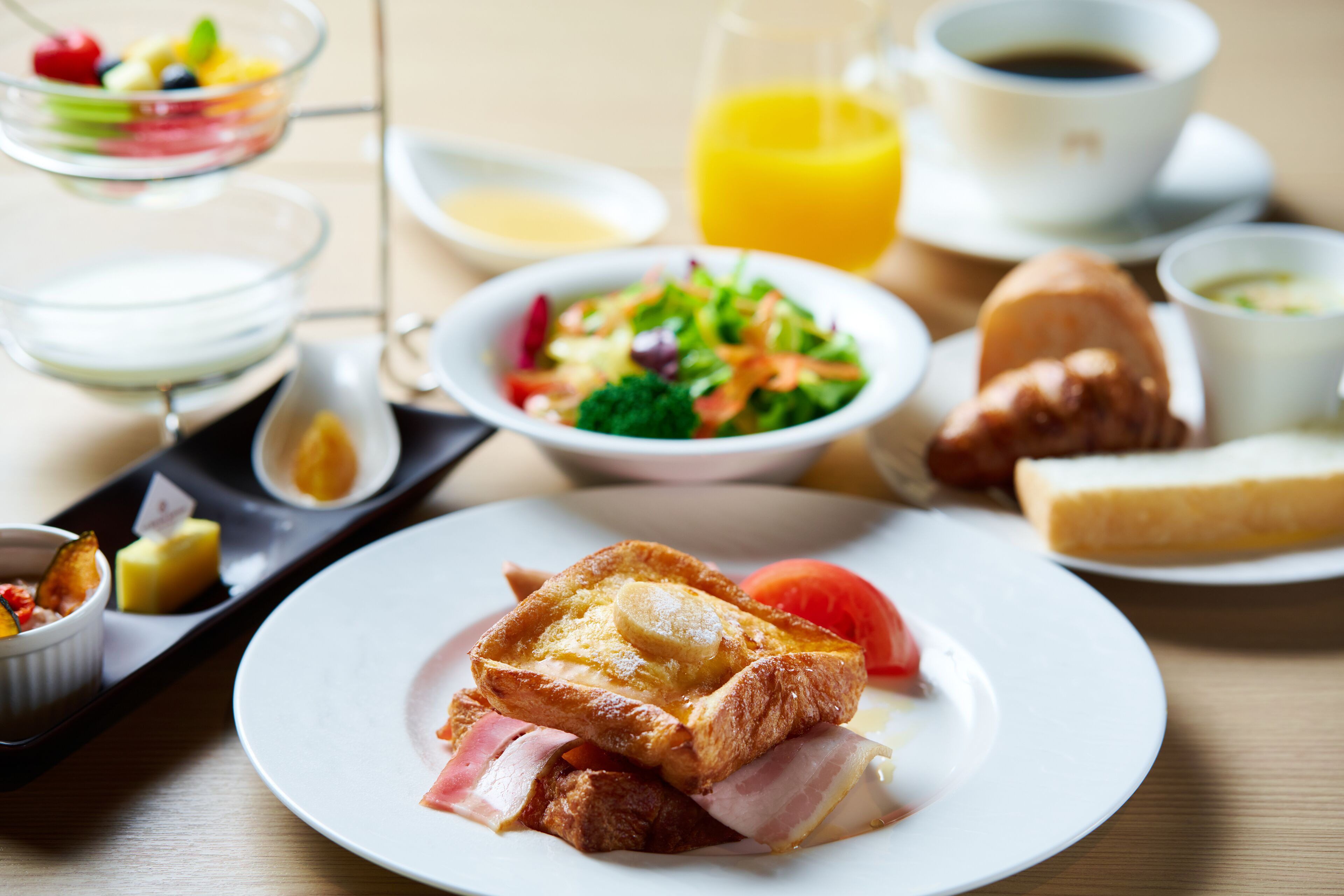 Una mesa de desayuno con tostadas, jamón y huevos en un plato, un tazón de ensalada, un croissant y un vaso de jugo de naranja.