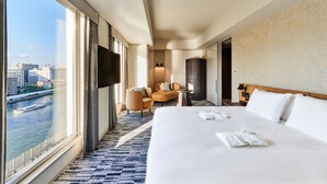 Habitación de hotel con una cama grande, zona de estar con sillas, televisión y vista al río.