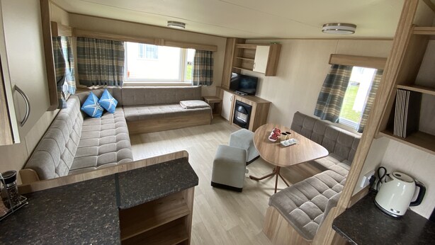 A compact RV interior with a sofa, a dining table, a TV, and a kitchenette. - 3 Bed 8 Berth Caravan in California Cliffs - D53 (Great Yarmouth)