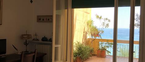 Ein Schlafzimmer mit einem Bett, einem Schreibtisch und einem Stuhl. Eine Chandelierleuchter und Blick auf das Meer durch eine offene Tür.