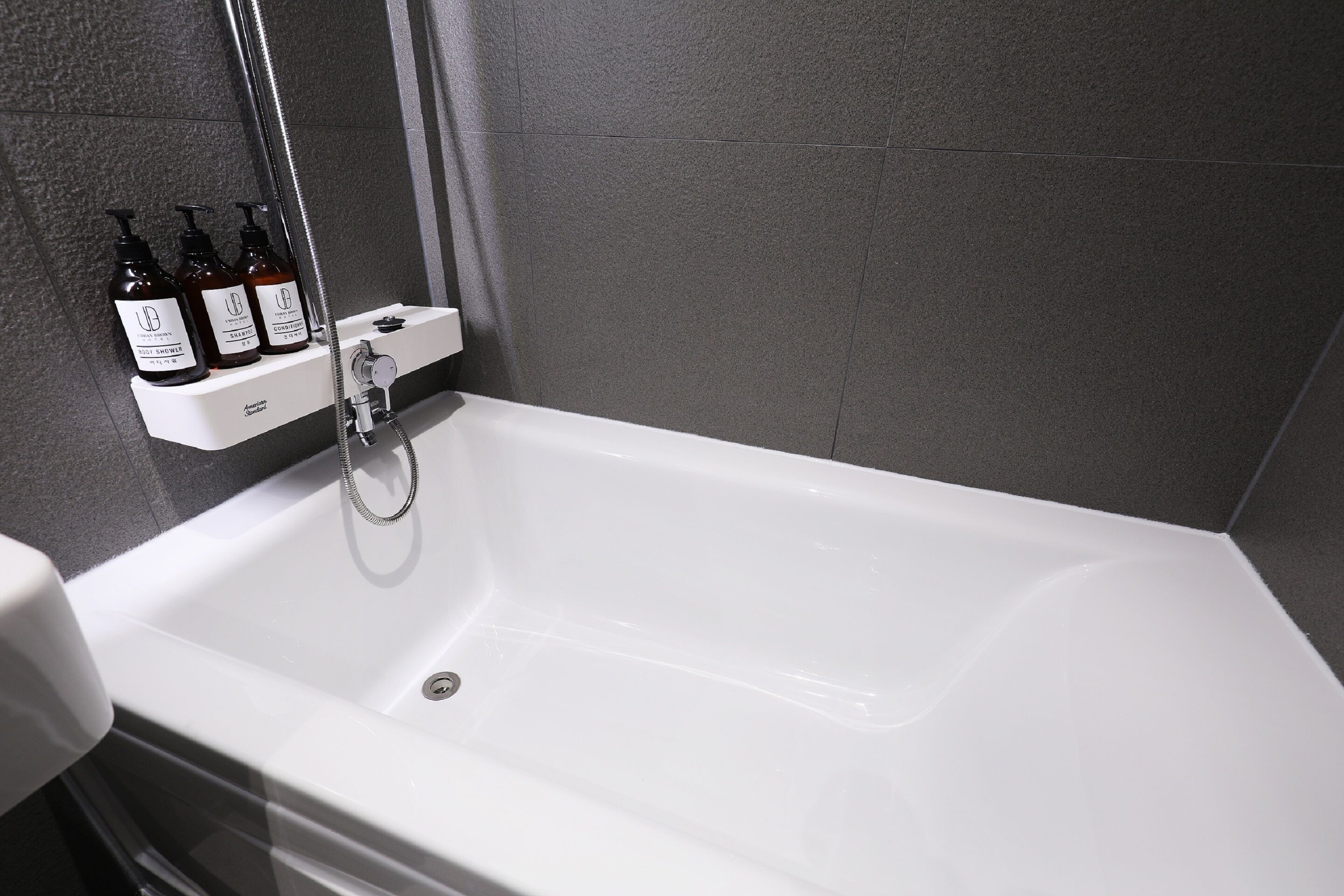 A white bathtub with a handheld showerhead, a white sink, and three bottles of shampoo on a shelf.