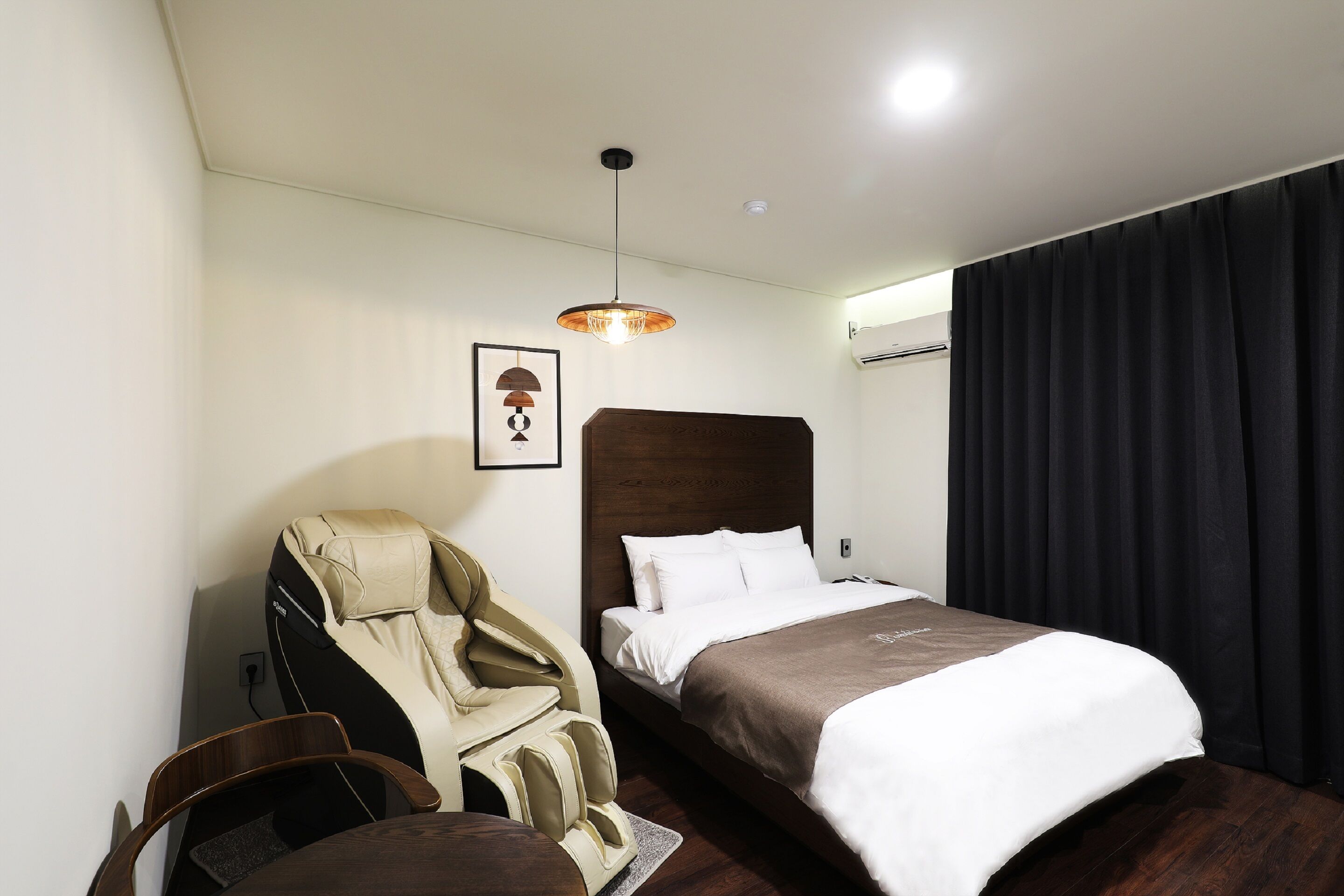 A hotel room with a bed, a massage chair, a desk, and a pendant light.