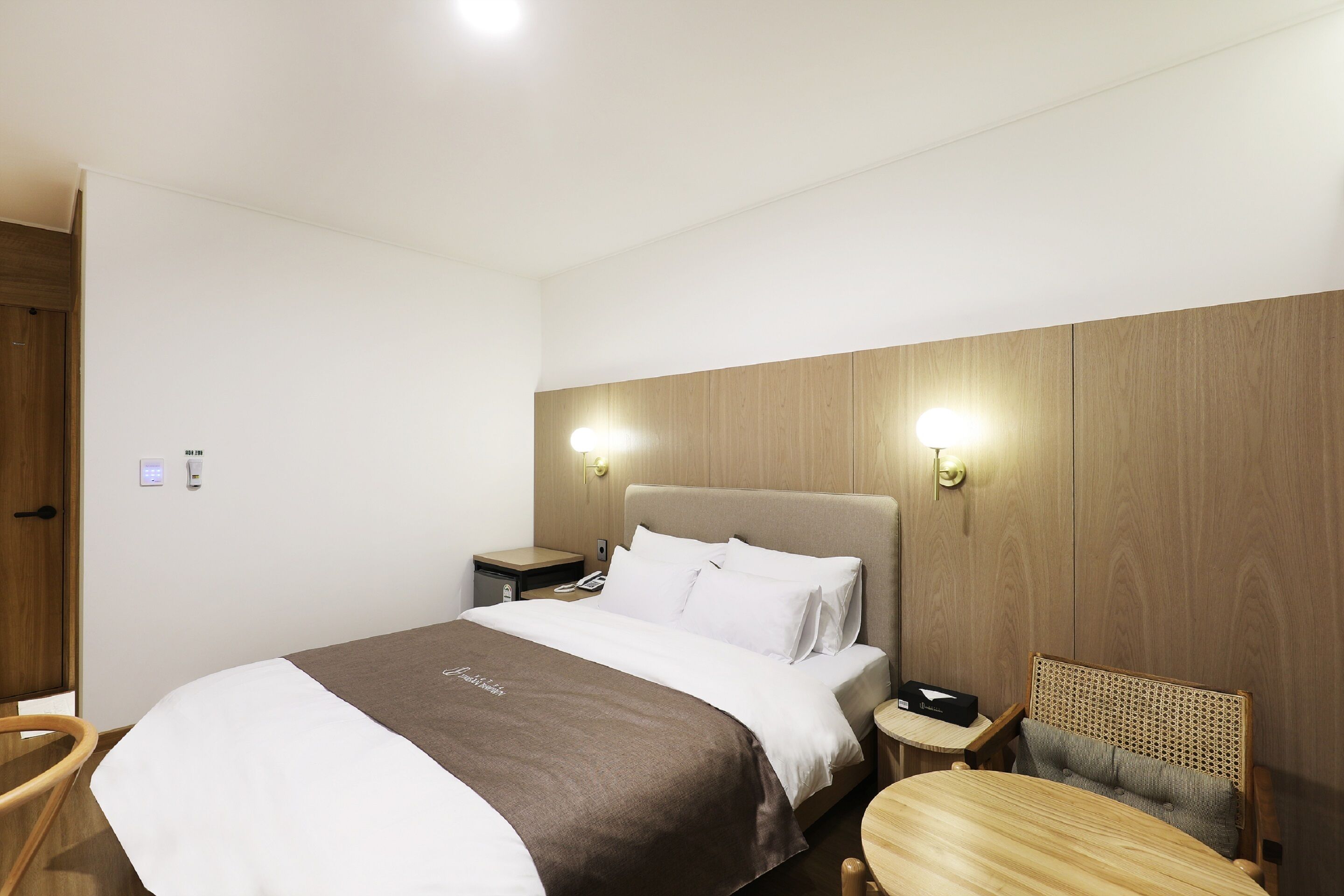 Habitació d'hotel amb un llit gran, una taula de nit amb una làmpada, una cadira i un panell de fusta al capçal.