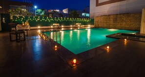 A swimming pool with a stone wall and a table with chairs, surrounded by string lights. - Rosewood Apartment Hotel - Pantnagar (Kichha)
