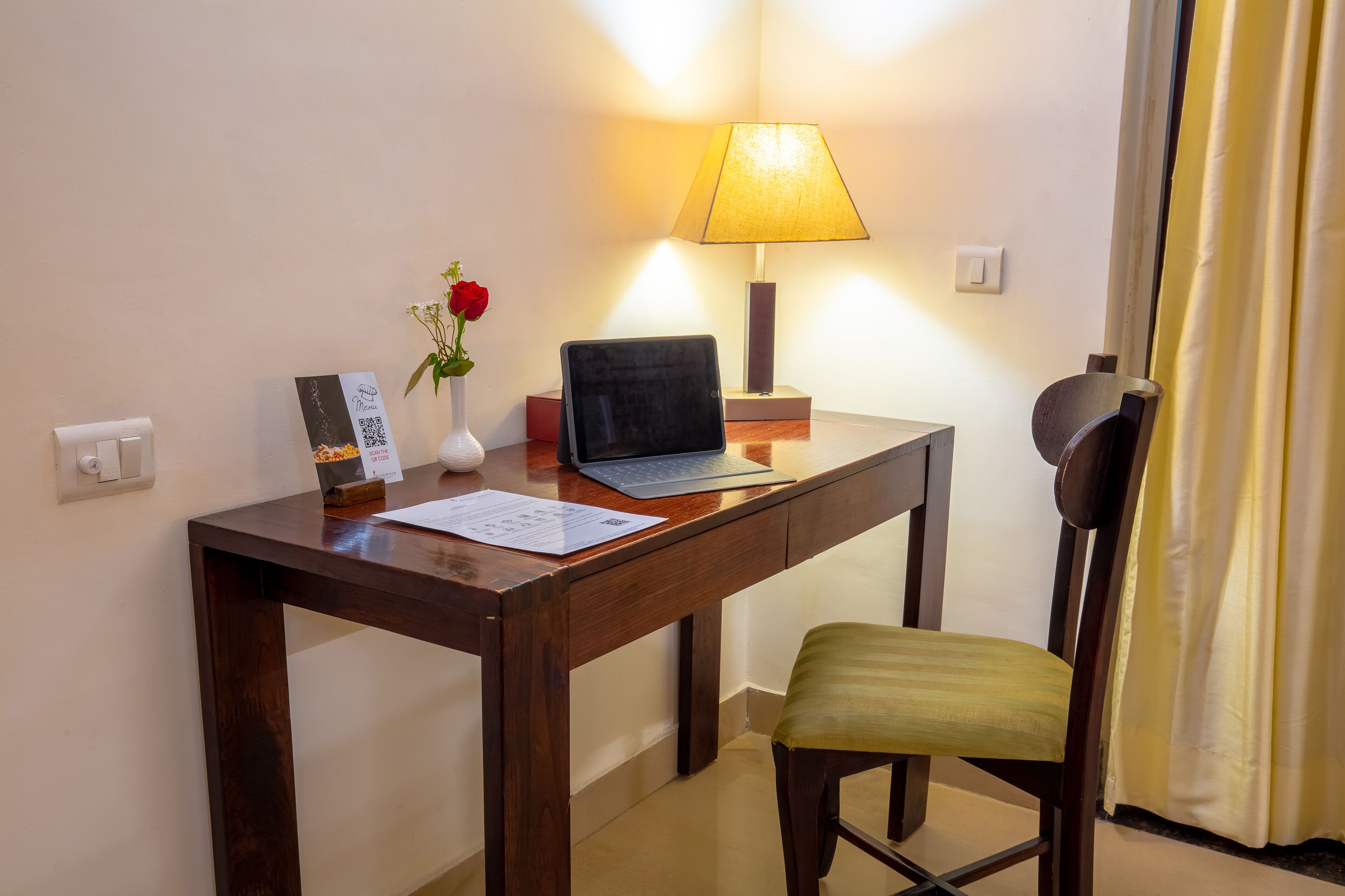 Un bureau en bois avec un ordinateur portable, un vase contenant une fleur rouge et une lampe.
