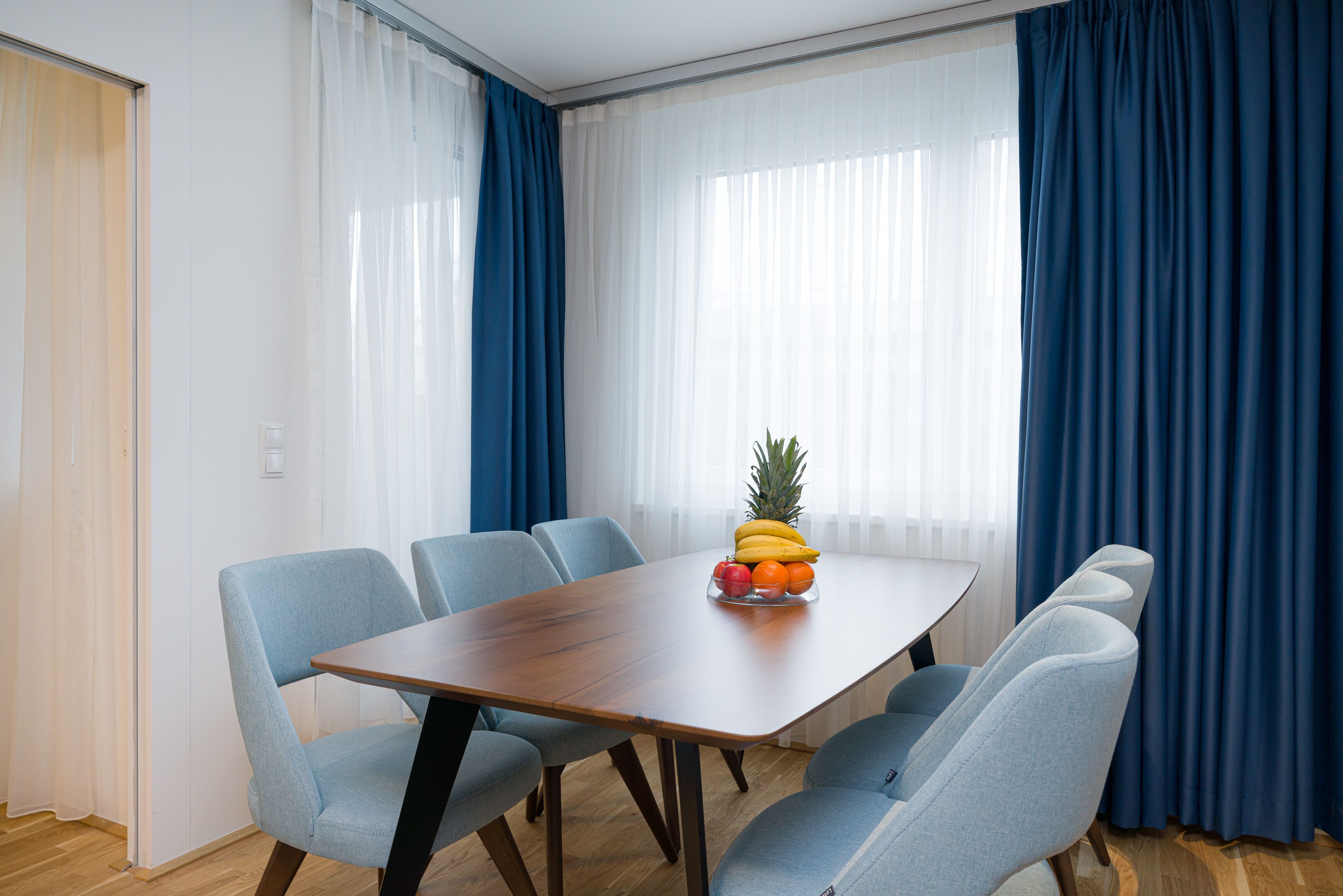 Uma área de refeições com uma mesa de madeira e seis cadeiras azuis. Uma fruteira com bananas, maçãs e laranjas está sobre a mesa.